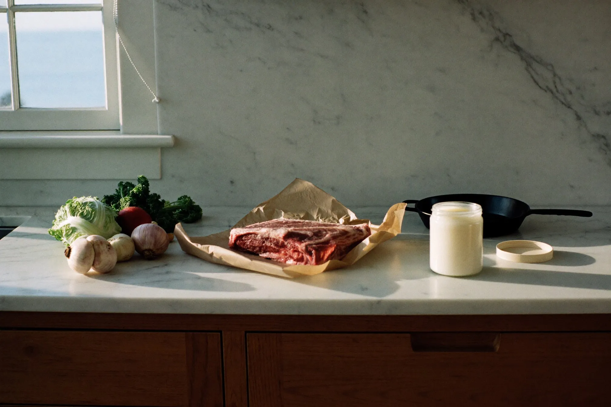 An open jar of tallow, a cast-iron pan, vegetables and steak on a kitchen counter.